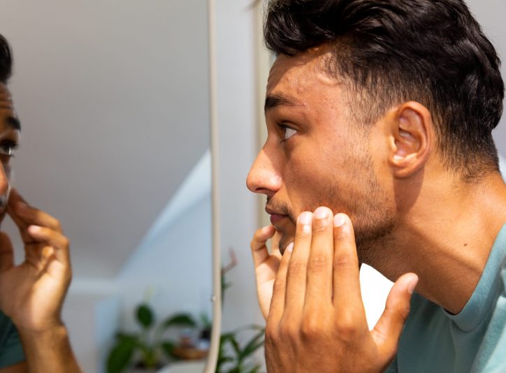 Biracial man touching his face, looking at himself in bathroom mirror. Lifestyle, self care and domestic life, unaltered.